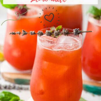 Three glasses of bright red strawberry lavender lemonade are garnished with lavender sprigs and surrounded by basil leaves, with the text “Homemade Strawberry Lavender Lemonade” and “thetastesoflife.com” on the image.
