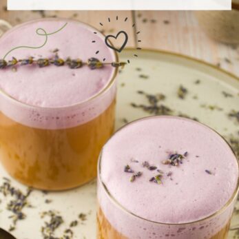 Two glass mugs filled with creamy lavender lattes topped with purple foam and garnished with dried lavender buds, sitting on a tray scattered with lavender flowers. Text reads “Creamy Lavender Latte” and “thetasteoflife.com”.
