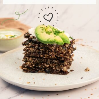 A stack of seed crackers topped with avocado slices and herbs sits on a light plate. Scattered seeds and dip are nearby. Text reads “Healthy Seed Crackers” and “thetastesoflife.com.”.