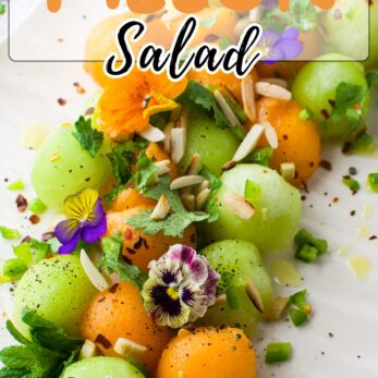 Colorful summer melon salad with cantaloupe and honeydew balls, garnished with mint leaves, edible flowers, sliced almonds, and spices, on a white surface. Text overlays read Summer Melon Salad and get the recipe.