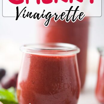 A glass jar filled with bright red roasted cherry vinaigrette sits on a table, with another jar blurred in the background. Text on the image reads Roasted Cherry Vinaigrette and get the recipe, with a website link.