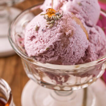 Two scoops of Honey Lavender Frozen Yogurt in a glass dish, topped with lavender buds and being drizzled with honey, with a blurred background and a jar of honey nearby.