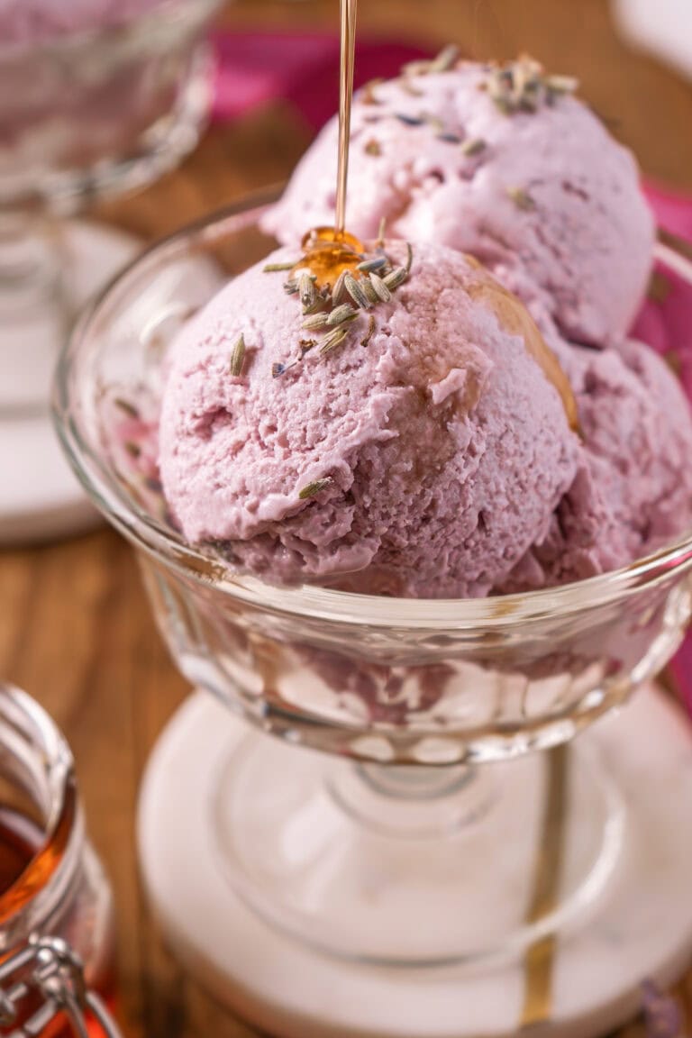 Two scoops of Honey Lavender Frozen Yogurt in a glass dish, topped with lavender buds and being drizzled with honey, with a blurred background and a jar of honey nearby.