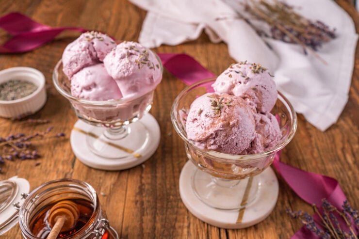 Two glass dishes filled with scoops of Honey Lavender Frozen Yogurt sit on a wooden table, surrounded by ribbons, dried lavender, a small bowl of herbs, a jar of honey, and a white cloth.