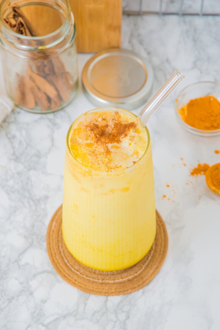 A tall glass of iced golden milk with a glass straw, topped with a sprinkle of cinnamon. The drink is creamy and vibrant yellow, sitting on a cork coaster on a marble surface. In the background are a jar of cinnamon sticks, a small bowl of turmeric powder,