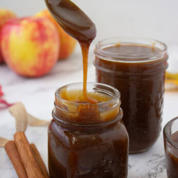 A spoonful of dark caramel sauce drizzled into a glass jar, with cinnamon sticks, star anise, and apples nearby.