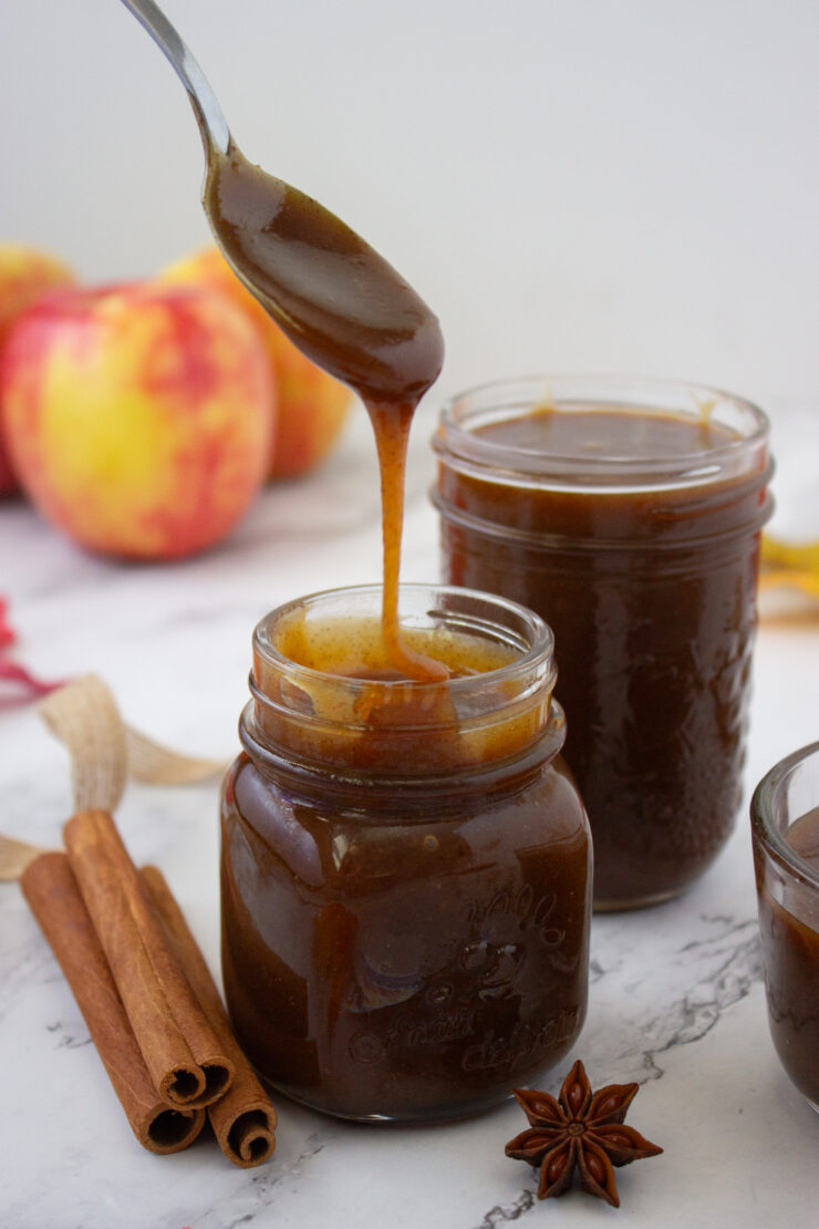 A spoonful of dark caramel sauce drizzled into a glass jar, with cinnamon sticks, star anise, and apples nearby.