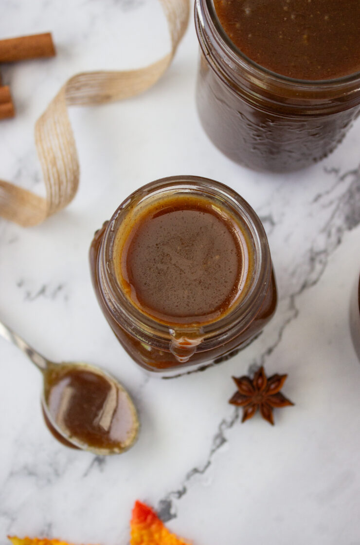 Top-down view of an open glass jar of caramel sauce with a spoon, cinnamon sticks, and a star anise on a marble surface.