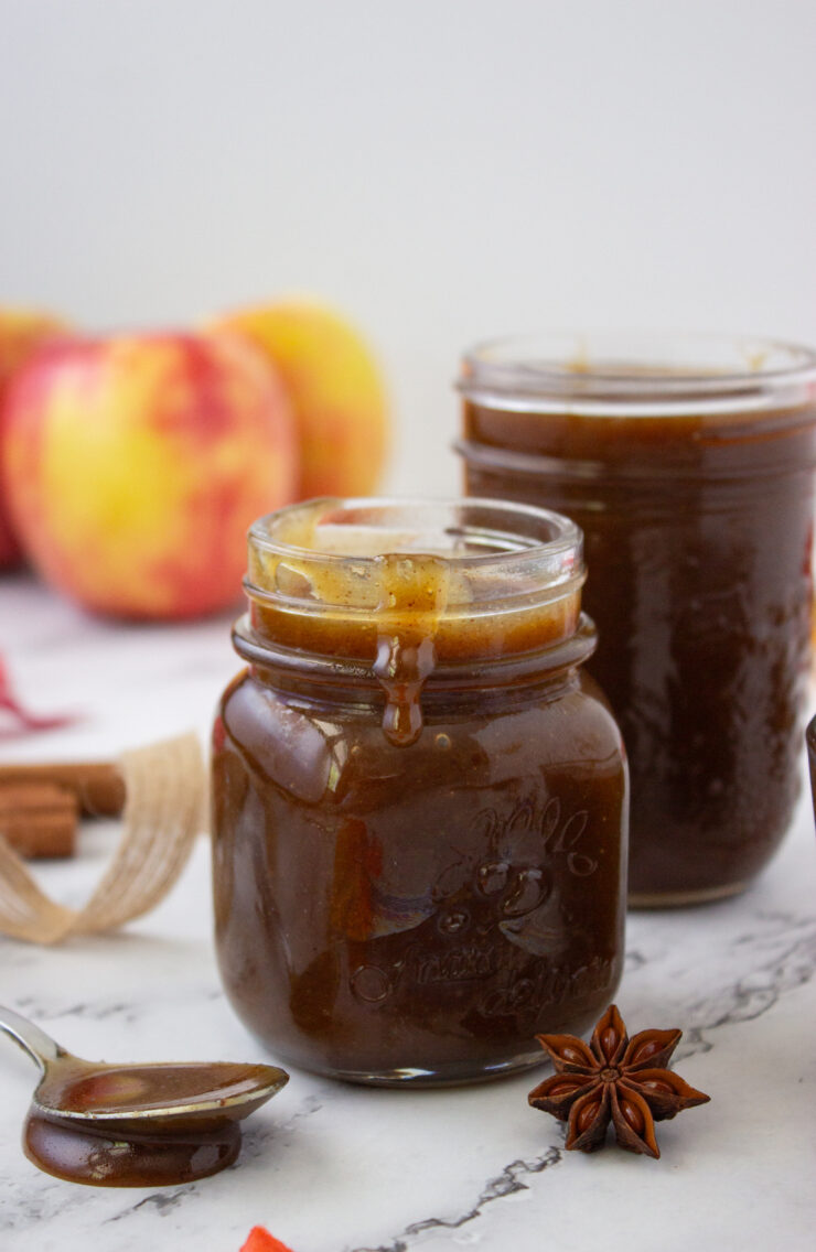Two small glass jars filled with dark apple butter, accompanied by a spoon, a star anise, and fresh apples in the background.