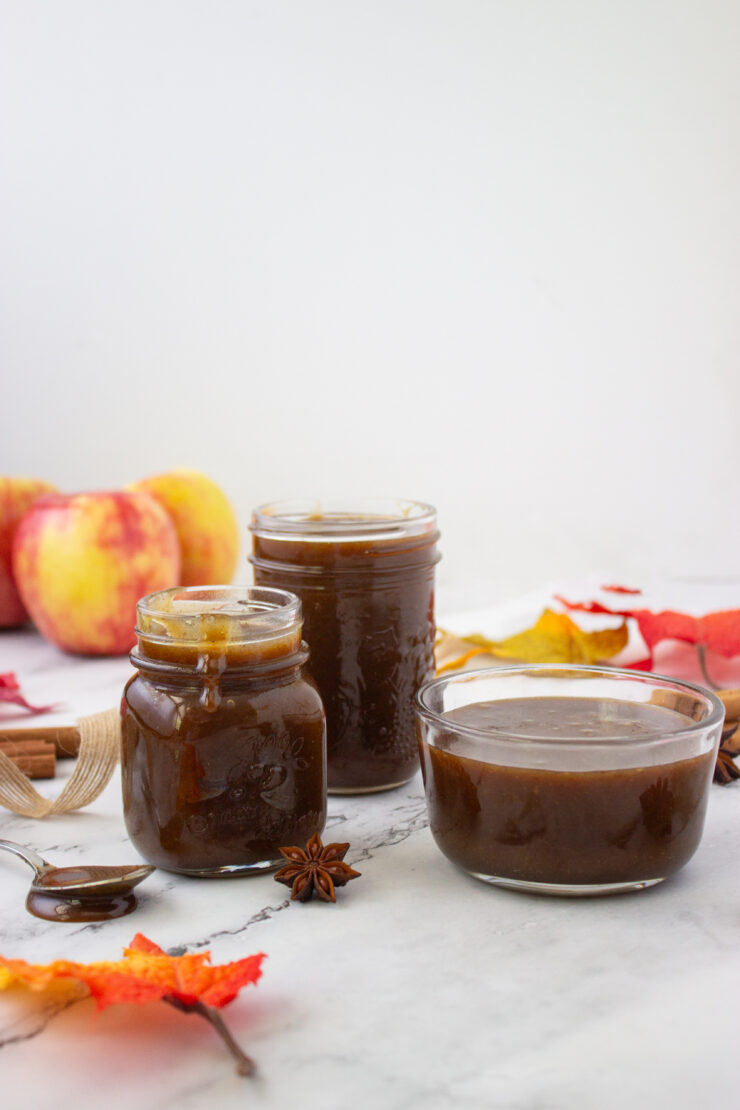 Two glass jars and a small bowl filled with dark apple butter, arranged on a marble surface with apples, star anise, and autumn leaves.