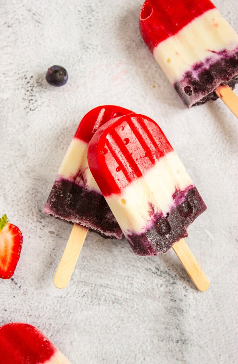 Two layered 4th of July Popsicles with red, white, and purple sections rest on a light surface. One is stacked on another, with a blueberry and sliced strawberry nearby. The popsicles appear fruity and refreshing.