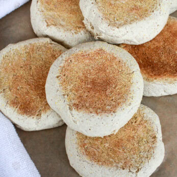 A close-up of several freshly baked English muffins with golden brown tops, arranged on parchment paper next to a white textured cloth.