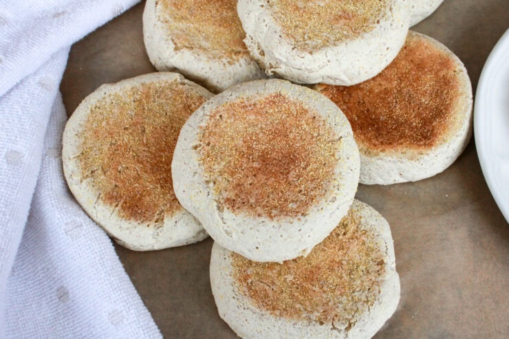 A close-up of several freshly baked English muffins with golden brown tops, arranged on parchment paper next to a white textured cloth.