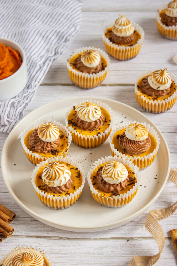 A plate of six cupcakes with chocolate and vanilla swirled frosting sits on a white wooden table, surrounded by more cupcakes, a bowl of orange puree, a striped cloth, and cinnamon sticks.