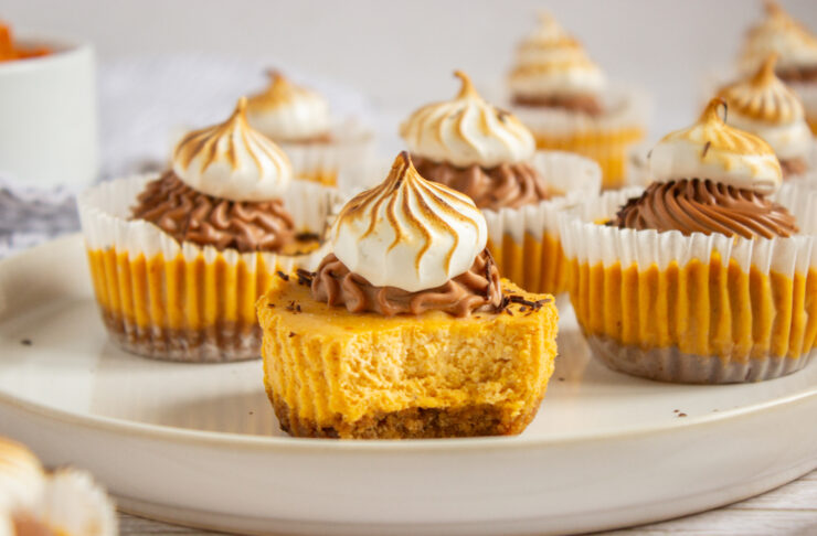 Mini pumpkin cheesecakes with whipped cream and chocolate frosting, displayed on a white plate. The cheesecake in front has a bite taken out, showing its creamy texture and crumbly crust.