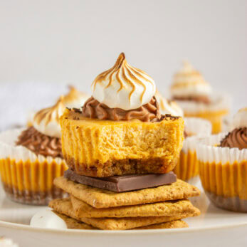 A Mini Pumpkin S'more Cheesecakes with a bite taken out, topped with swirled toasted meringue, sits on graham crackers and chocolate, surrounded by similar cupcakes on a plate.