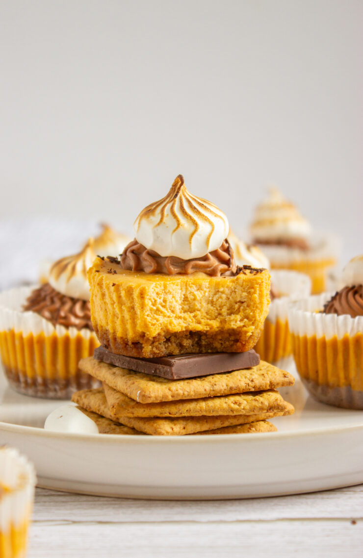 A smores cupcake with a bite taken out, topped with swirled toasted meringue, sits on graham crackers and chocolate, surrounded by similar cupcakes on a plate.