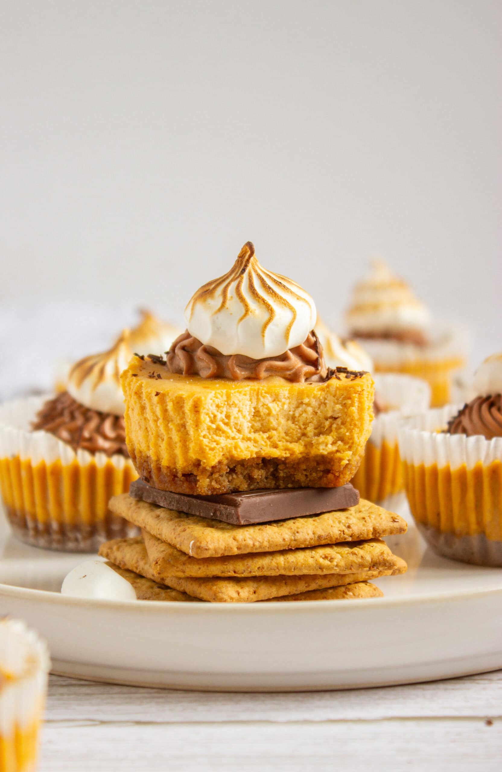 A Mini Pumpkin S'more Cheesecakes with a bite taken out, topped with swirled toasted meringue, sits on graham crackers and chocolate, surrounded by similar cupcakes on a plate.