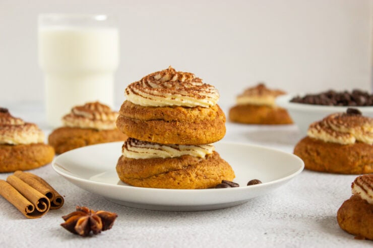 Two stacked cookies topped with swirls of frosting and cocoa powder on a white plate, with cinnamon sticks, star anise, and coffee beans nearby.