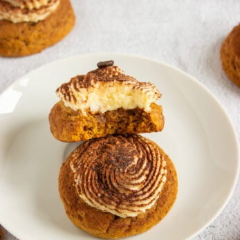 Tiramisu-flavored cookies topped with swirled frosting and cocoa powder on a white plate, one cookie broken in half.