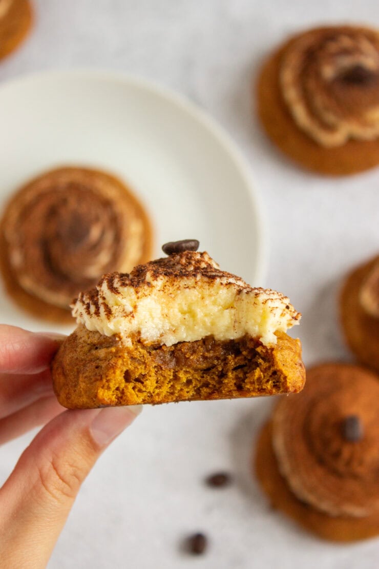 A hand holds a bitten pumpkin spice cookie topped with cream cheese frosting, cocoa powder, and a single coffee bean.