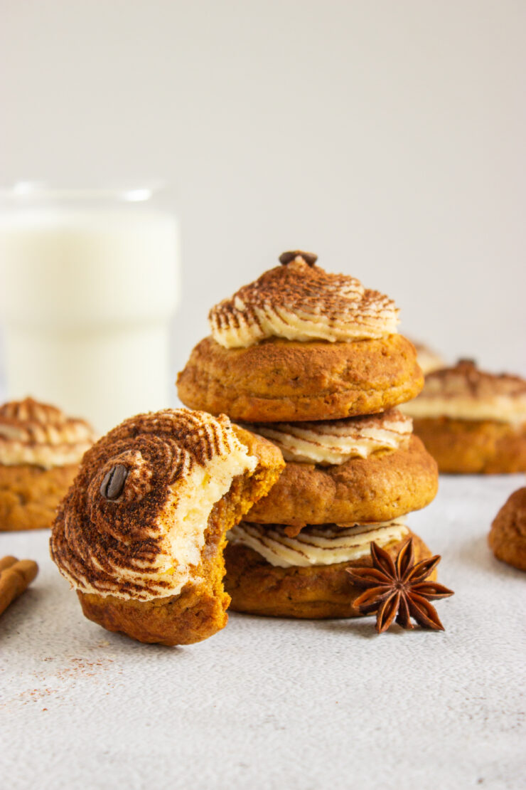 A stack of three Pumpkin Tiramisu Cookies (Gluten-Free) topped with cream frosting and a coffee bean, with a partially eaten cookie in the foreground and a glass of milk in the background.