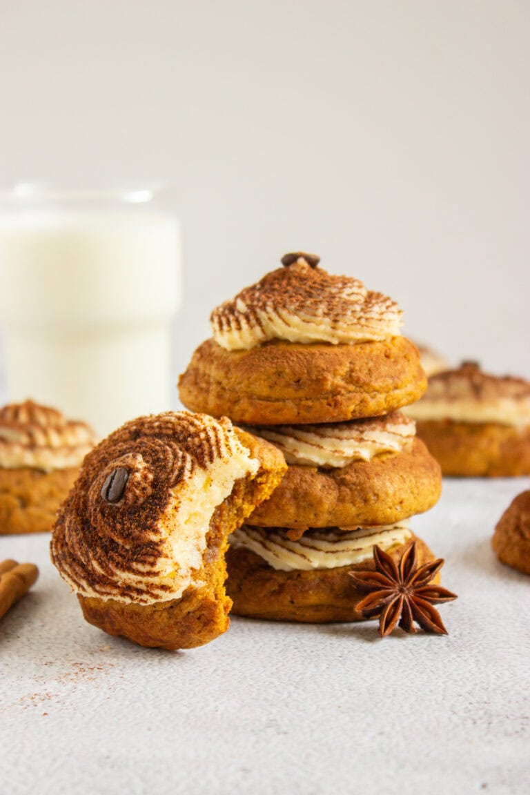 A stack of three Pumpkin Tiramisu Cookies (Gluten-Free) topped with cream frosting and a coffee bean, with a partially eaten cookie in the foreground and a glass of milk in the background.