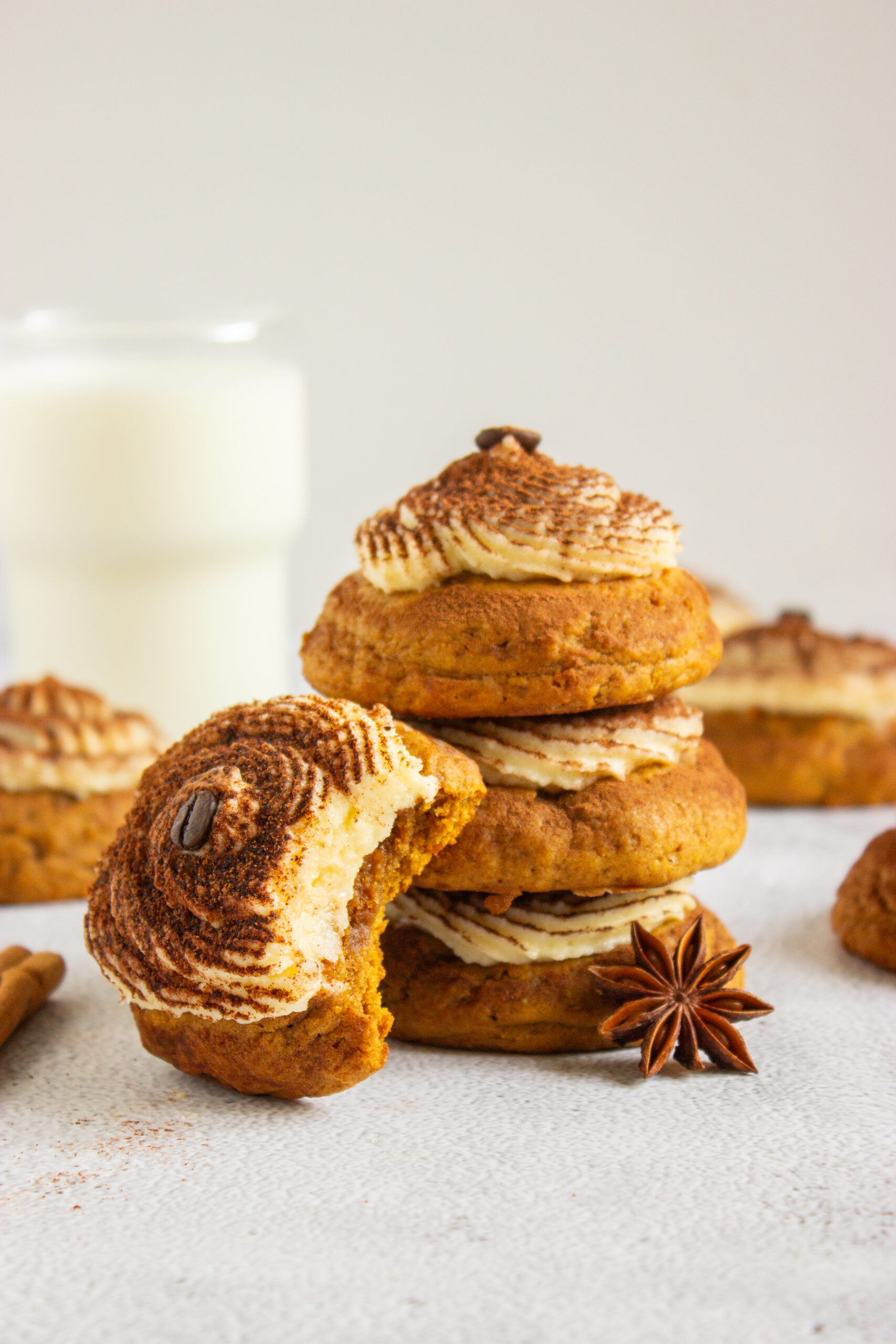 A stack of three Pumpkin Tiramisu Cookies (Gluten-Free) topped with cream frosting and a coffee bean, with a partially eaten cookie in the foreground and a glass of milk in the background.