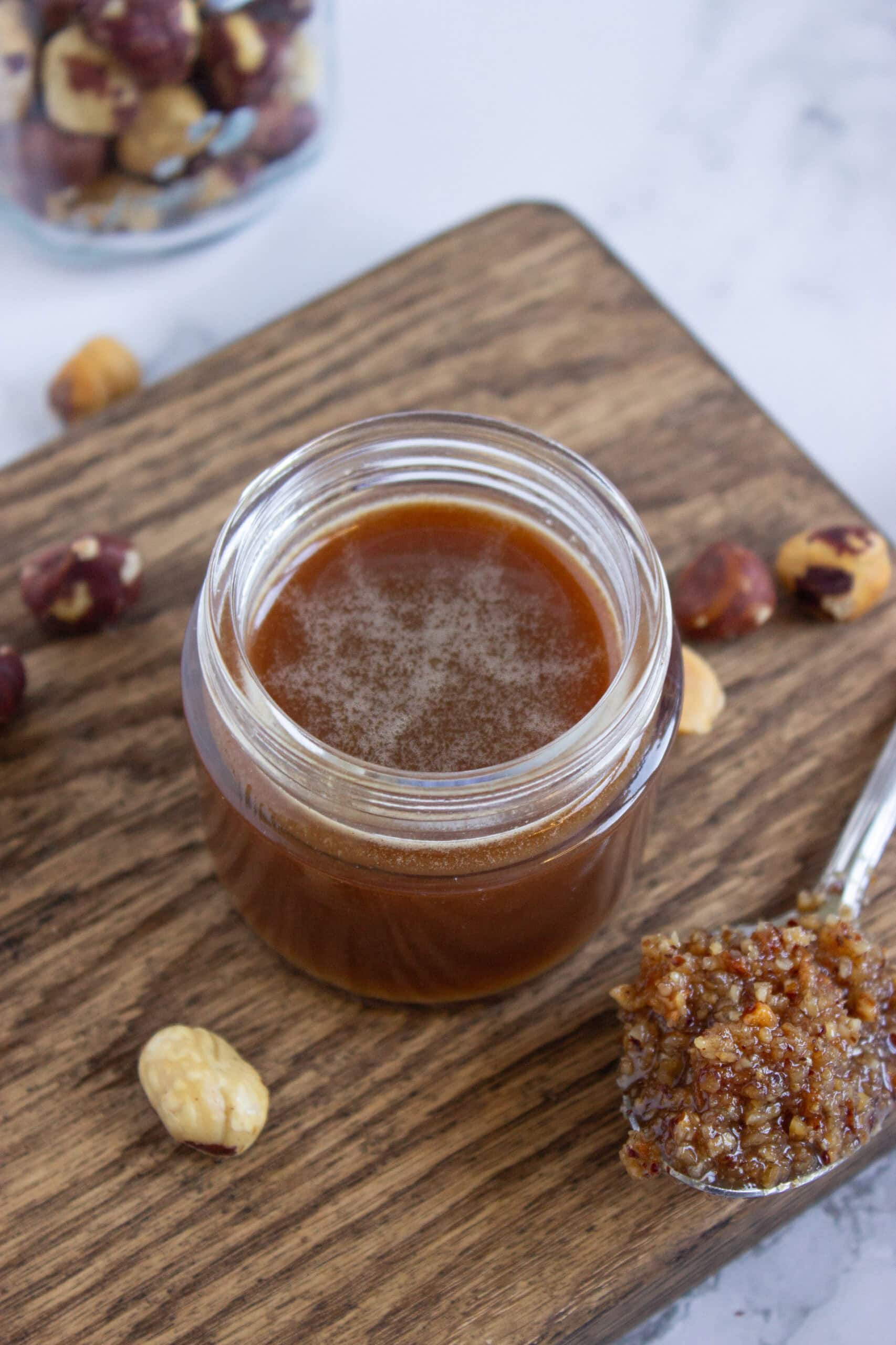 A small glass jar filled with hazelnut butter sits on a wooden board, surrounded by whole hazelnuts. A spoon with a chunky mixture rests beside the jar. A jar of hazelnuts is visible in the background.