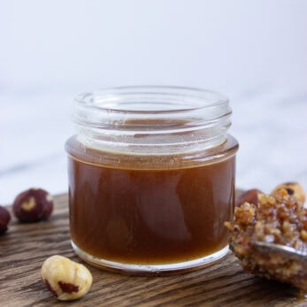 A small glass jar filled with brown hazelnut syrup sits on a wooden surface, with whole hazelnuts and a spoonful of syrup nearby. The background is softly blurred.