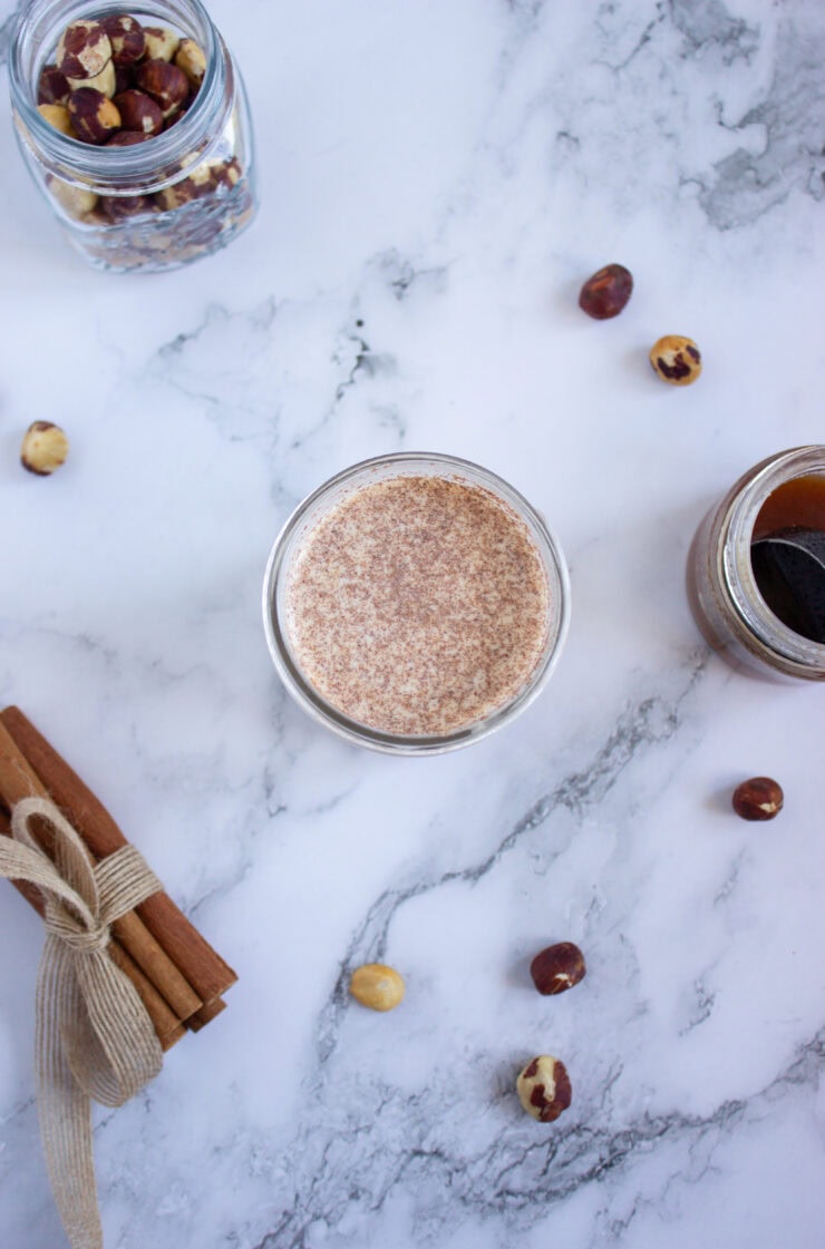 A glass of frothy beverage on a marble surface, surrounded by scattered hazelnuts, a jar of mixed nuts, a jar of dark liquid, and a bundle of cinnamon sticks tied with twine.