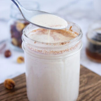 A Homemade Hazelnut Creamer spoon hovers over a glass jar filled with creamy white liquid, sprinkled with cinnamon. Hazelnuts and a glass jar are in the background on a wooden surface.