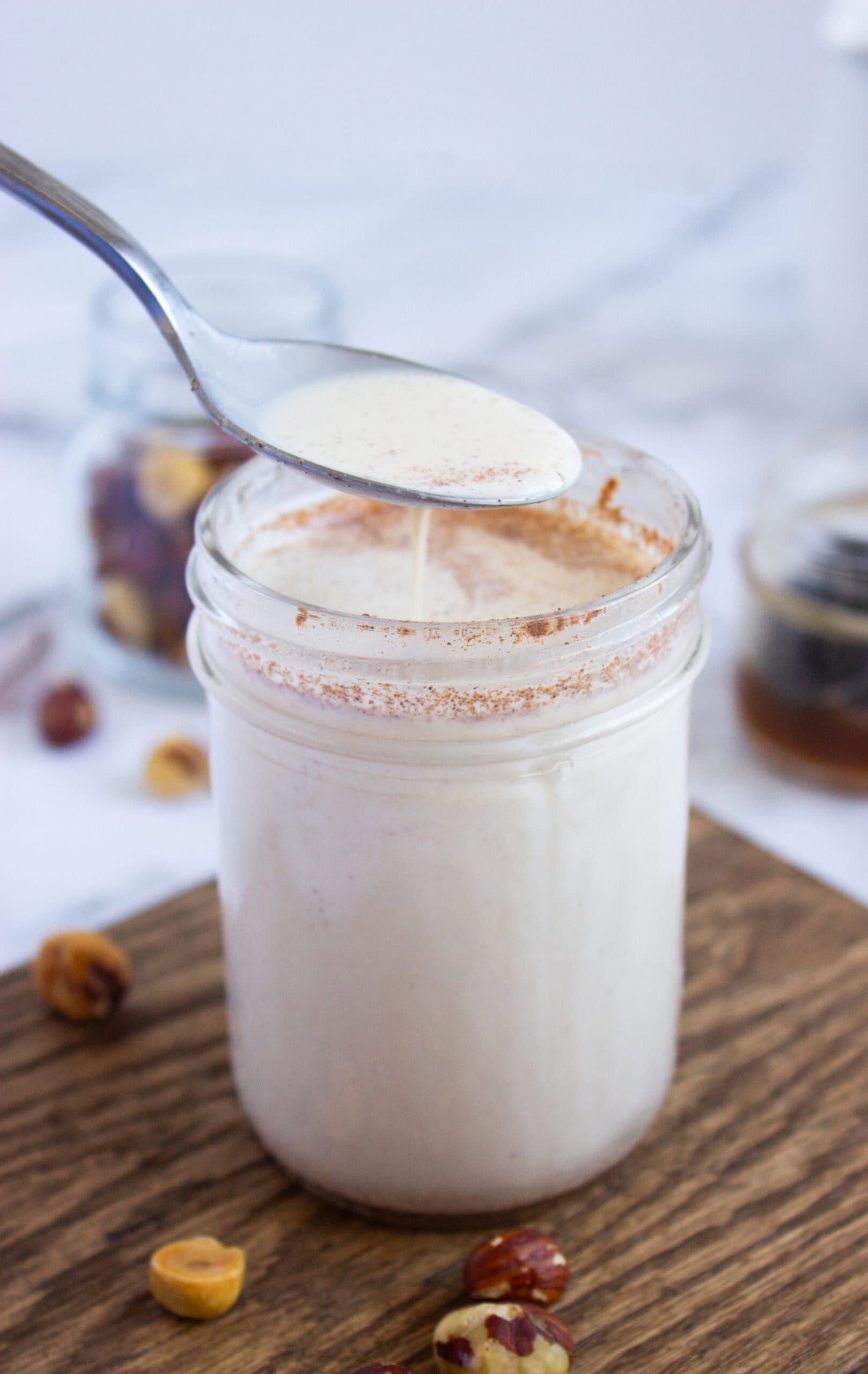 A Homemade Hazelnut Creamer spoon hovers over a glass jar filled with creamy white liquid, sprinkled with cinnamon. Hazelnuts and a glass jar are in the background on a wooden surface.
