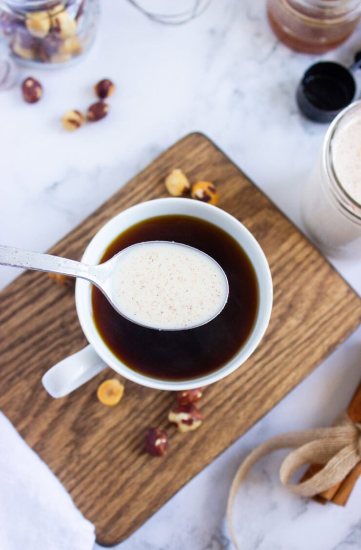 A spoonful of creamy liquid is held above a cup of black coffee on a wooden board, with scattered hazelnuts and a jar in the background on a white surface.