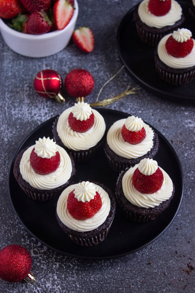 A black plate with six chocolate cupcakes topped with white frosting and whole strawberries, resembling Santa hats. Red Christmas ornaments and a bowl of strawberries are in the background.