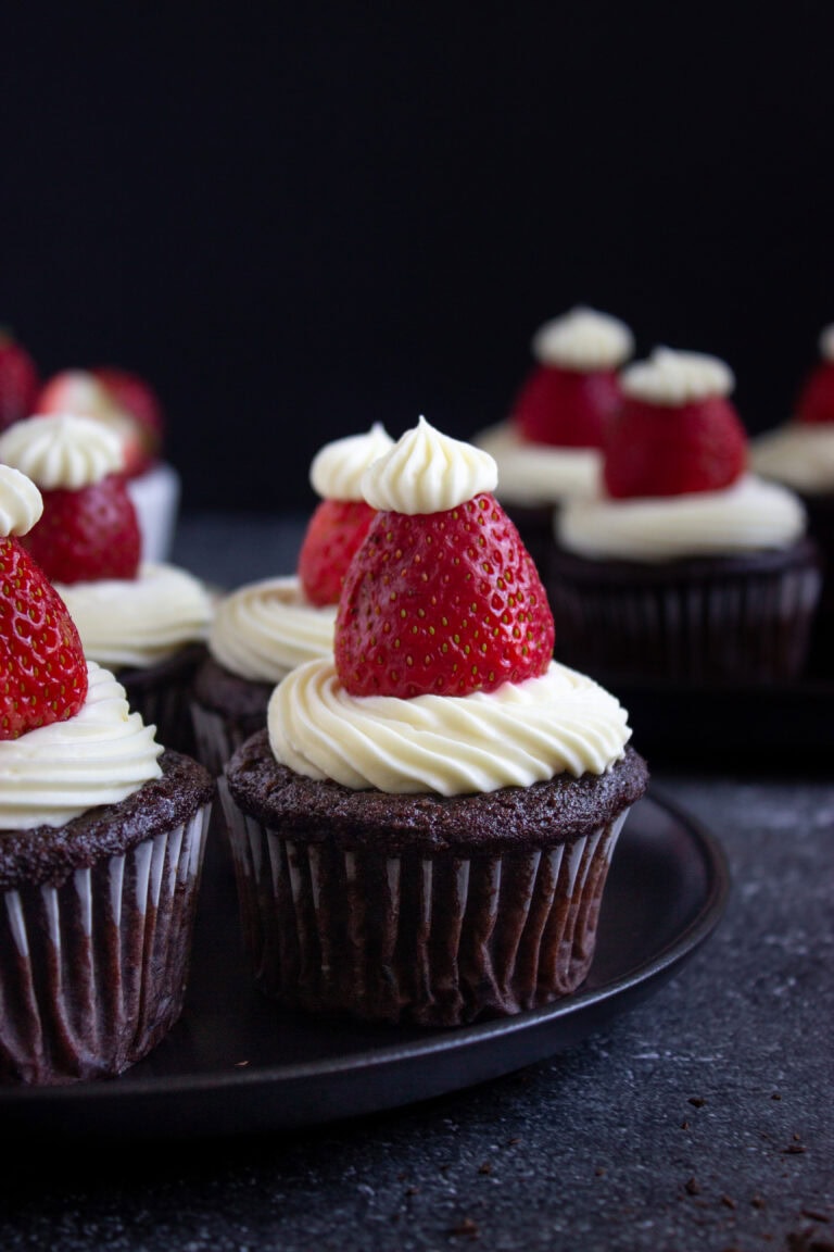 Santa Hat Chocolate Cupcakes (Gluten-Free) topped with swirls of white frosting and a fresh strawberry, finished with a small dollop of frosting on each strawberry, arranged on a dark plate against a dark background.