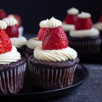 Santa Hat Chocolate Cupcakes (Gluten-Free), topped with swirls of white frosting and whole strawberries, arranged on black plates with a dark background.