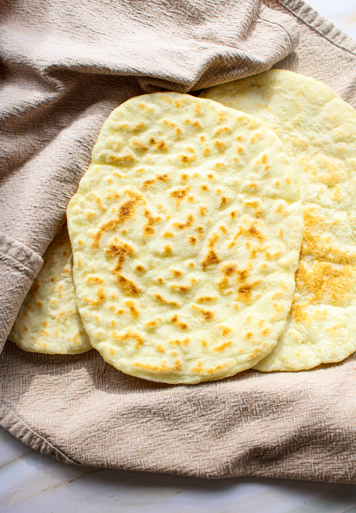 Three pieces of homemade Gluten-Free Naan Bread textured beige cloth on a light-colored surface.