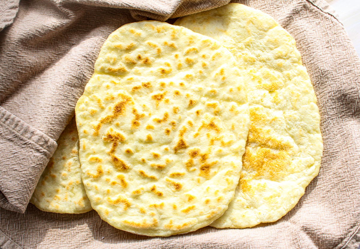 Three oval-shaped pieces of flatbread rest on a beige textured cloth, with golden brown spots indicating they are freshly cooked. The bread appears soft and slightly puffy.