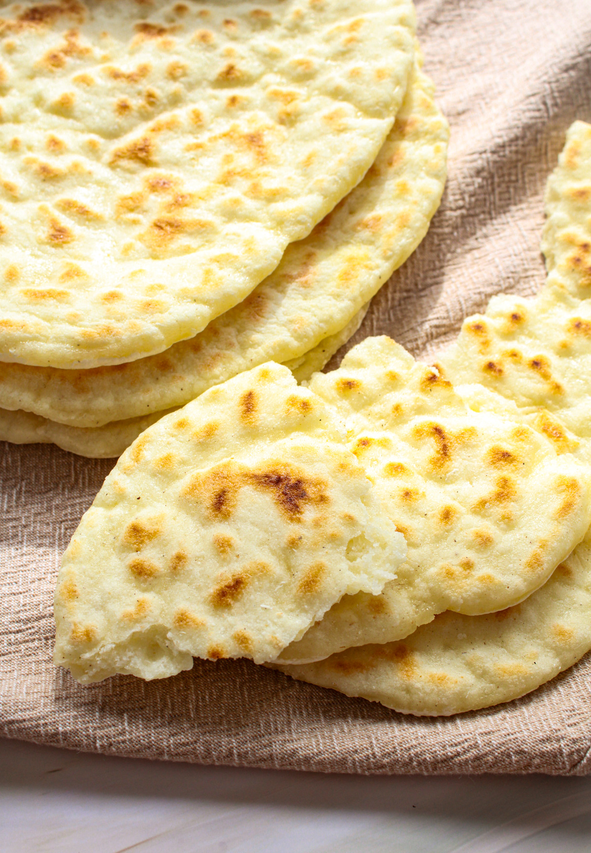 Several pieces of round, Gluten-Free Naan Bread are stacked and fanned out on a beige textured cloth, with one piece partially broken to show the soft interior.