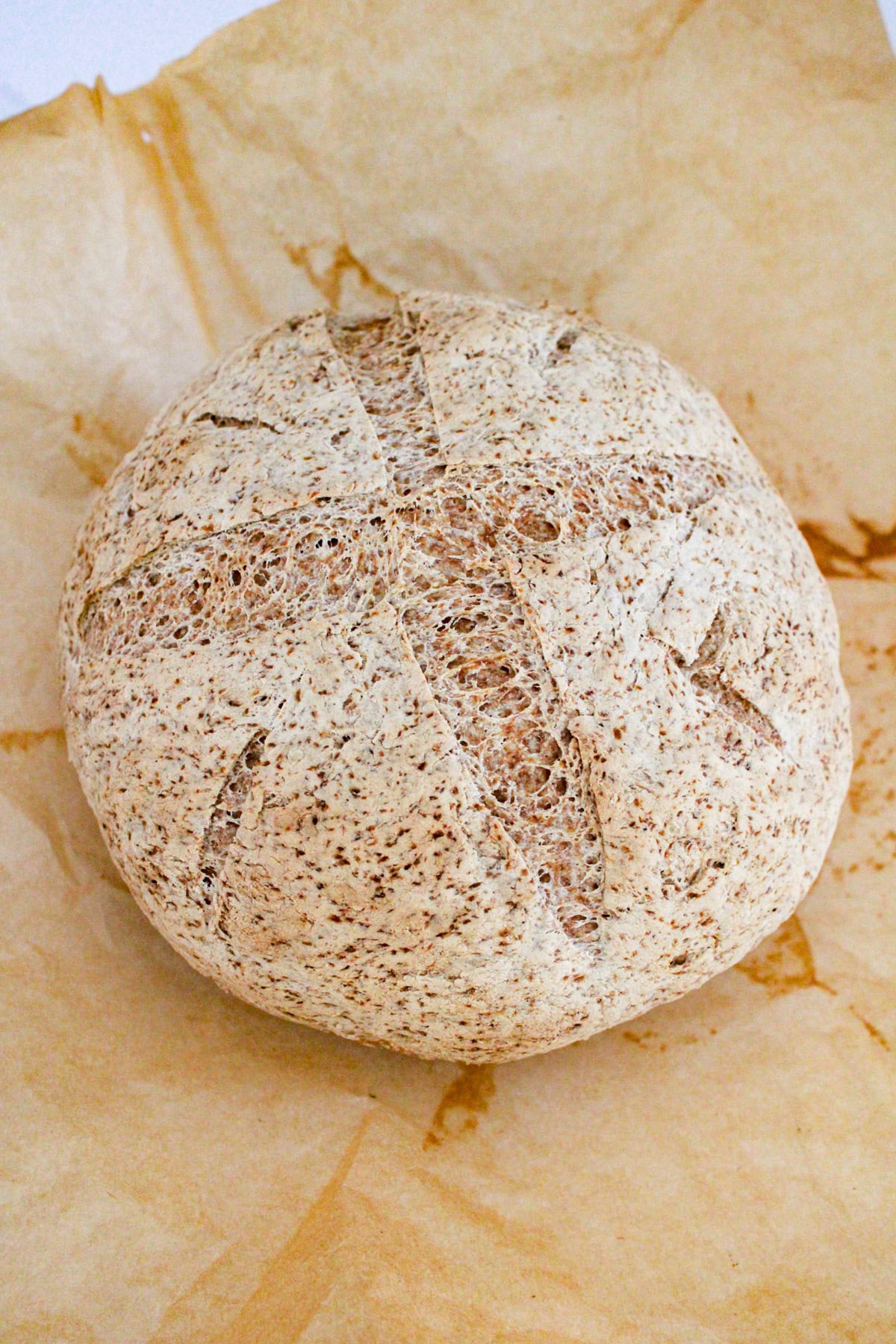 A round loaf of rustic bread with a textured, lightly browned crust sits on a sheet of parchment paper. The bread has deep scoring marks on top, forming a cross pattern.