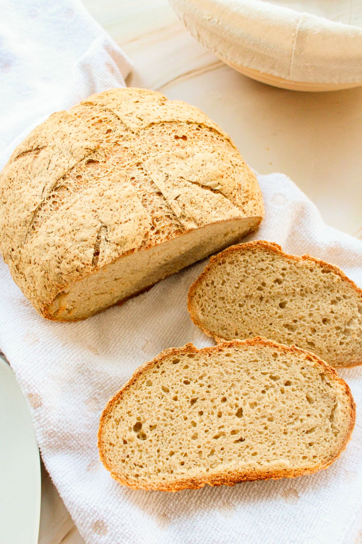 A round loaf of bread with a rustic crust sits on a white towel, with two slices cut and placed nearby, showing the breads airy, textured interior.