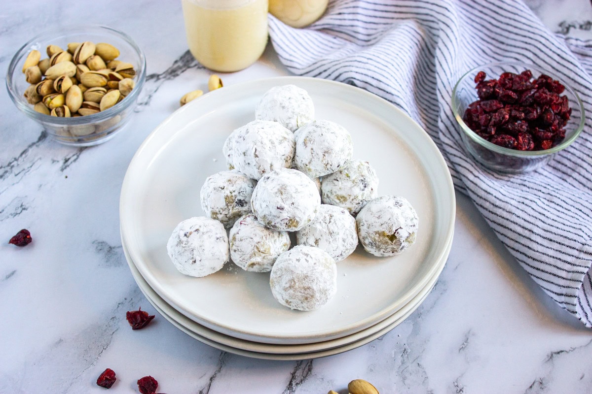 A stack of powdered sugar-coated dessert balls on a white plate, surrounded by bowls of pistachios and dried cranberries, with a striped cloth and drink in the background on a marble surface.