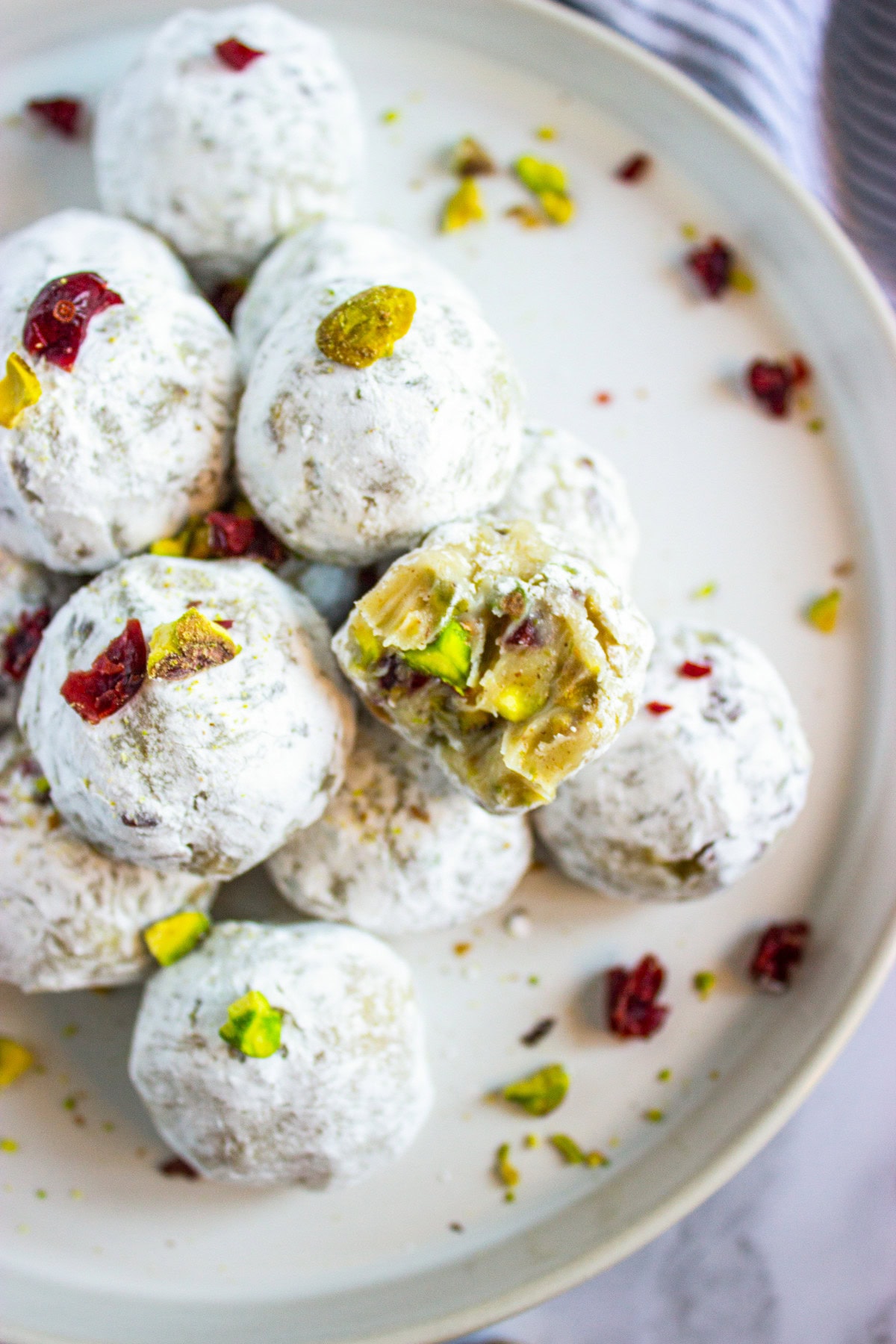A plate of Eggnog Pistachio Cranberry Truffles bite-sized balls, with one cut open to reveal a filling containing pistachios and dried cranberries. Bits of pistachio and cranberry are sprinkled on the plate.