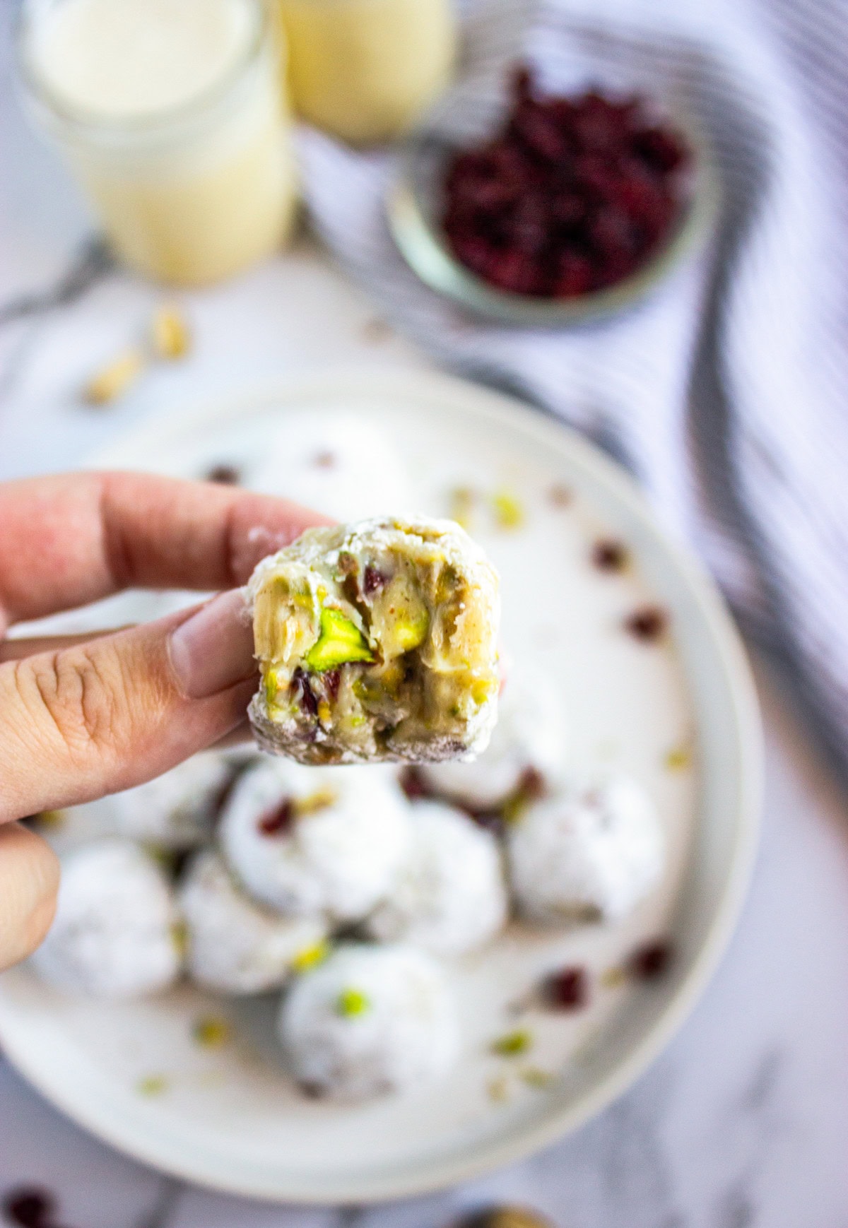 A hand holds up a round, powdered sugar-covered dessert with a bite taken out, revealing a filling with nuts and dried fruit. A plate of similar treats, a glass jar, and a bowl of dried cranberries are in the background.