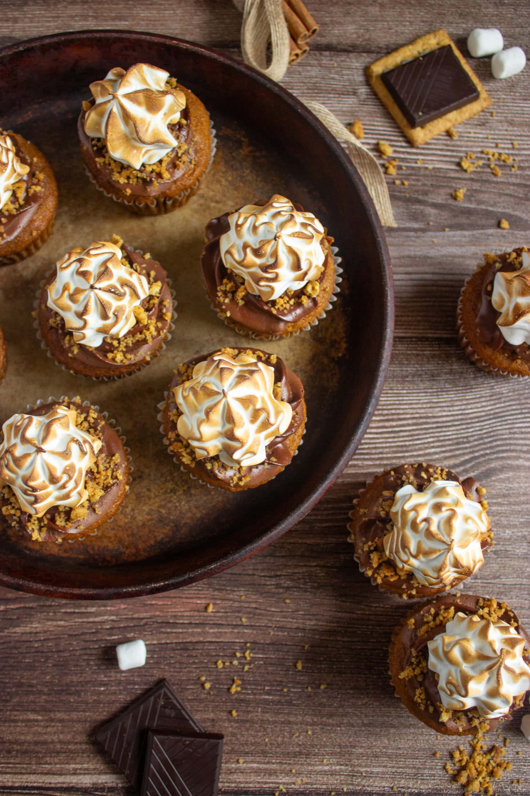 Angled shot of pumpkin s’mores cupcakes arranged in a round rustic pan, each topped with chocolate frosting, toasted meringue, and crumbled graham crackers