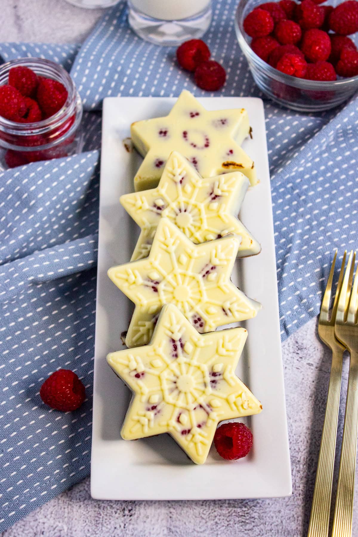 Star-shaped white chocolate desserts with raspberries are arranged on a rectangular white plate, surrounded by fresh raspberries in glass bowls and on a blue dotted cloth, with gold forks nearby.