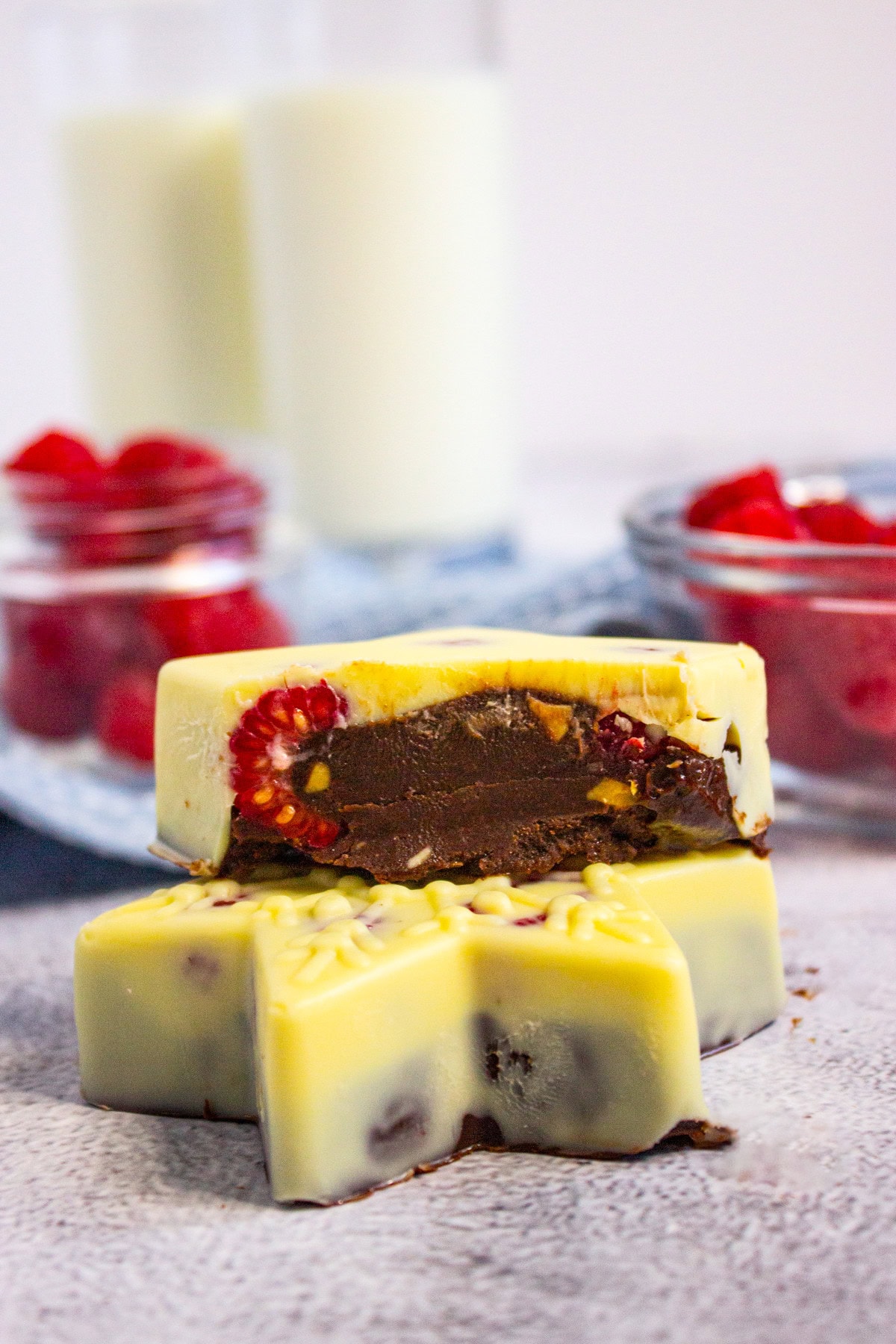 Two white chocolate star-shaped candies filled with chocolate and raspberries are stacked, with bowls of raspberries and two glasses of milk blurred in the background.