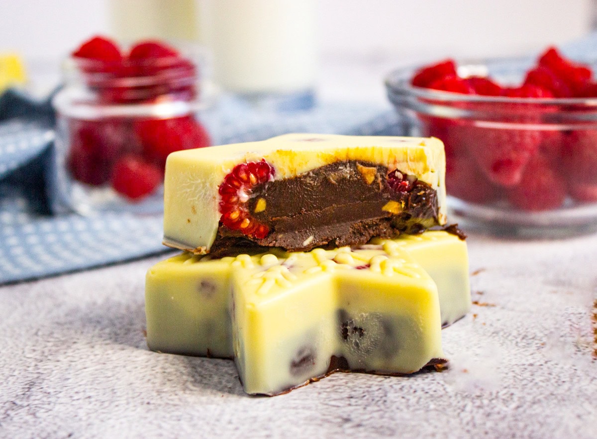 Two star-shaped white chocolate desserts with visible raspberries and dark chocolate filling are stacked. Bowls of fresh raspberries and a blue cloth are in the blurred background.