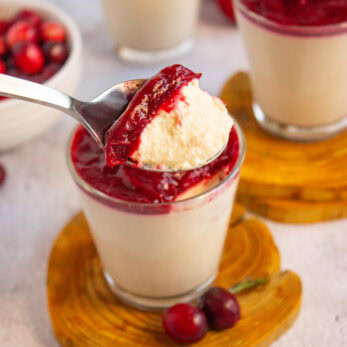 A spoonful of creamy Keto Cinnamon Panna Cotta with Cranberry Compote is held above a glass filled with the same dessert. Two more glasses and a bowl of cherries are in the background, all set on a light surface.